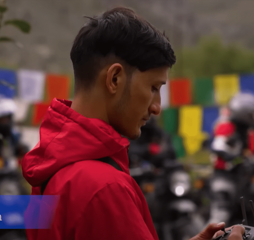 Concentrated male videographer in red adjusting camera settings with colorful prayer flags in soft focus in the background, depicting active outdoor videography.