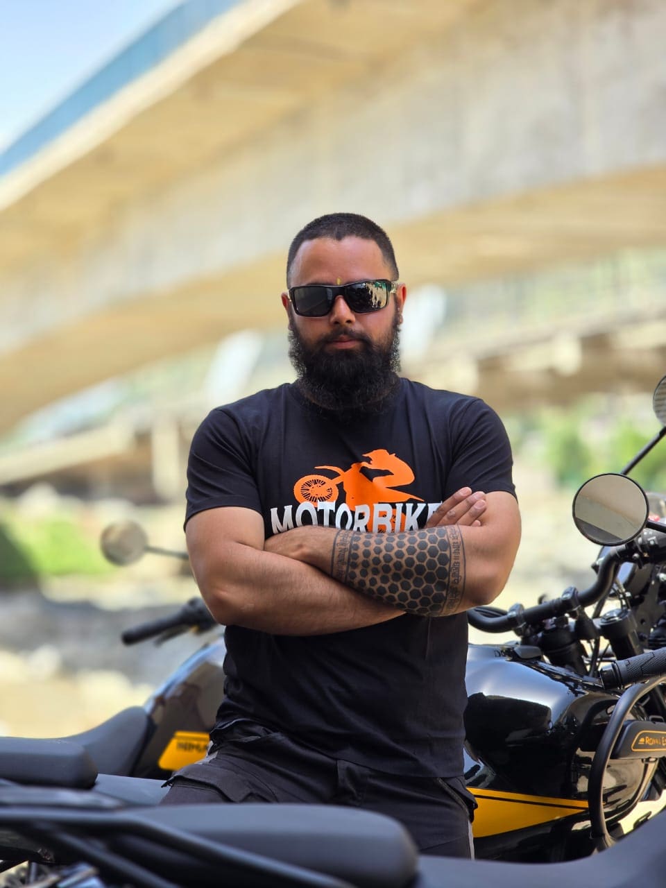 Akshay Thakur, founder of Motorbike Expeditions Ltd, wearing a black t-shirt and sunglasses, standing with arms crossed next to a Motorbike.