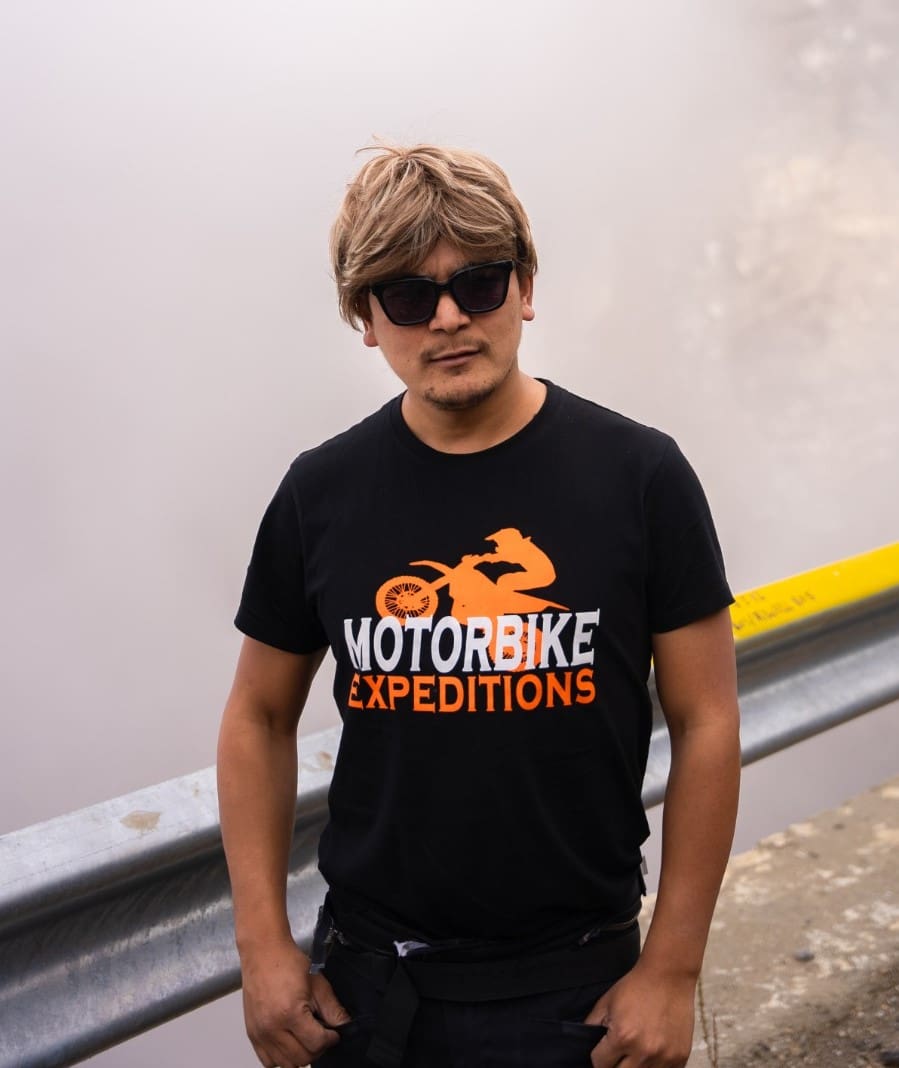 Deepak, a motorbike tour guide, wearing sunglasses and a black "Motorbike Expeditions" t-shirt, standing next to a metal railing on a foggy day.