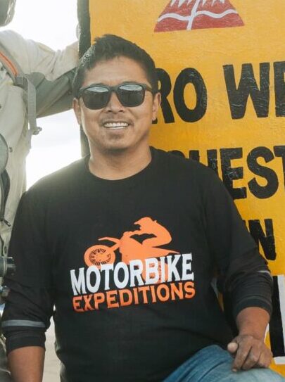 Gurmit, backup vehicle driver for Motorbike Expeditions, wearing sunglasses and a branded t-shirt in front of a signboard